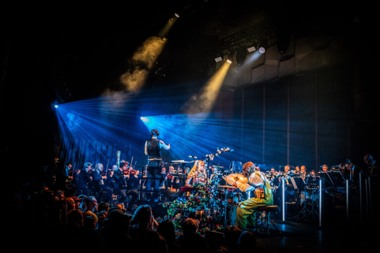 Een sfeervolle live muziekoptreden met een orkest en muzikanten op het podium, omgeven door kleurrijke belichting.