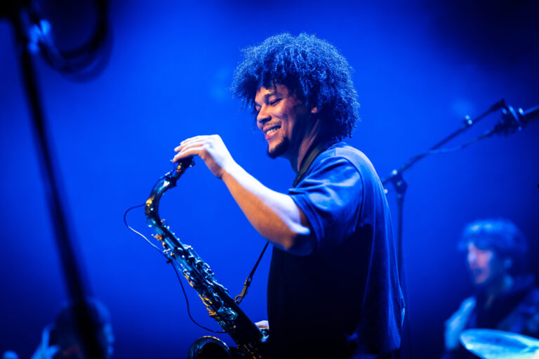 Een muzikant met krullend haar speelt saxofon op een podium met blauwe verlichting.