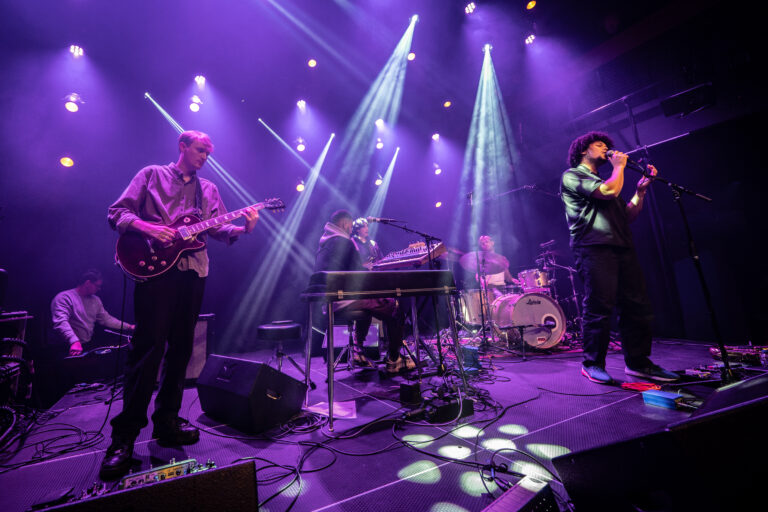 Een band speelt live op een podium, omringd door kleurrijke lichteffecten.