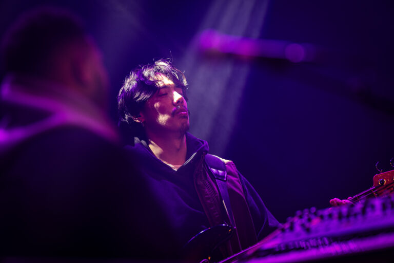 Een muzikant staat op het podium onder paarse lichten en kijkt de zaal in.