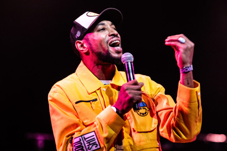 Een man in een felgele jas staat op het podium en zingt met een microfoon in zijn hand.