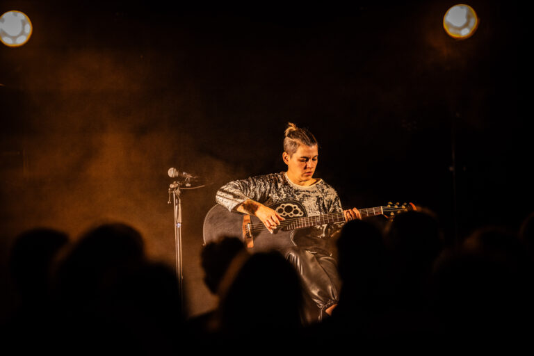 Een muzikante speelt gitaar op het podium terwijl een publiek in de schaduw toekijkt.