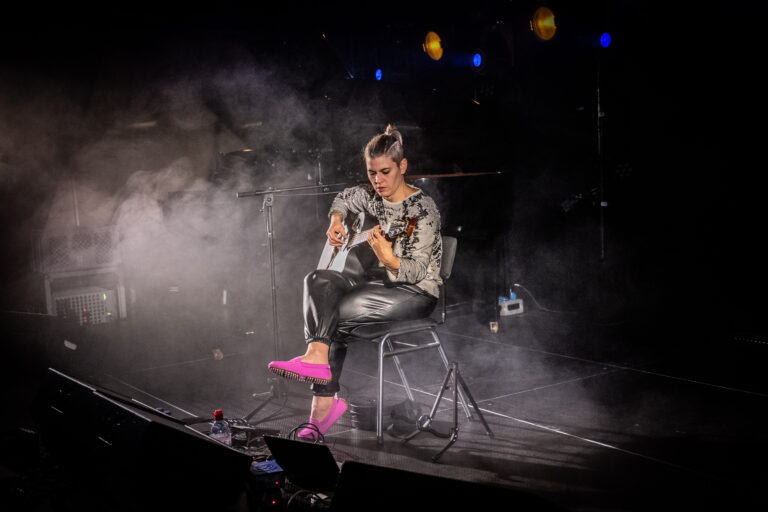 Een muzikante zit op een stoel in een mistige omgeving terwijl ze een gitaar bespeelt.