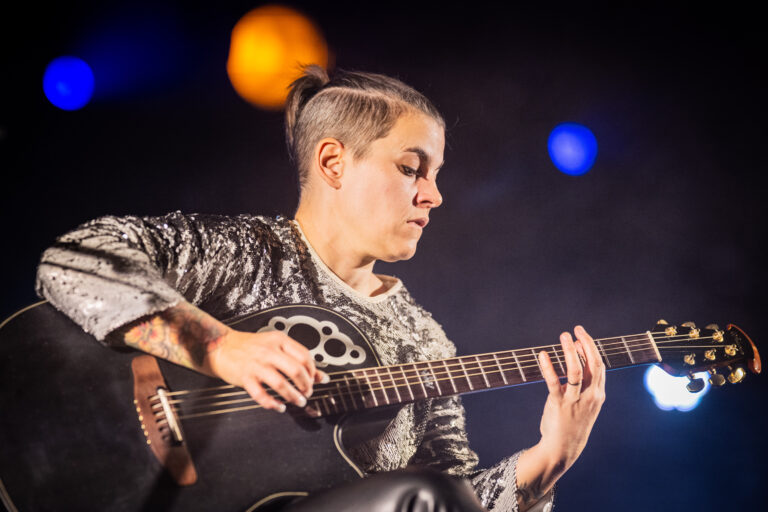 Een vrouw speelt gitaar op een podium, omringd door sfeerlichten.