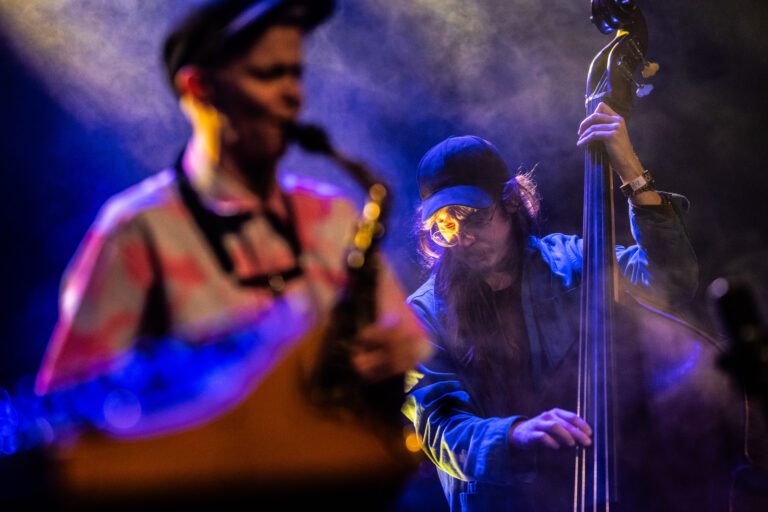 Een muzikant speelt saxophone terwijl een andere muzikant op de contrabas in de achtergrond staat, omringd door rook en kleurrijke verlichting.