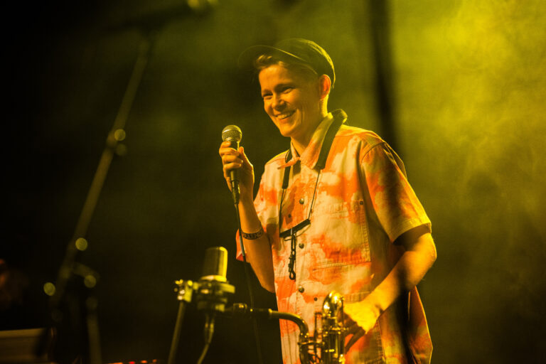 Een vrolijke muzikant met een microphone staat op het podium in een geel verlichte omgeving.