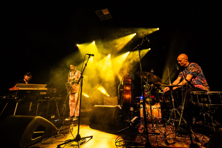 Een muzikant staat op een podium met een saxophone, omringd door rook en gele lichtstralen, terwijl een drummer naast hem speelt.