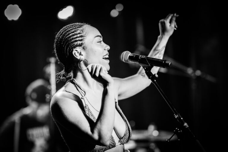 Een zangeres met vlechten staat in de schijnwerpers en zingt enthousiast in een microfoon.