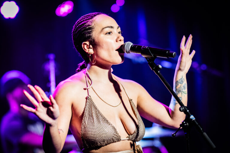 Een zangeres performt op een podium met een microfoon in haar hand, terwijl ze wordt verlicht door kleurrijke lichten.