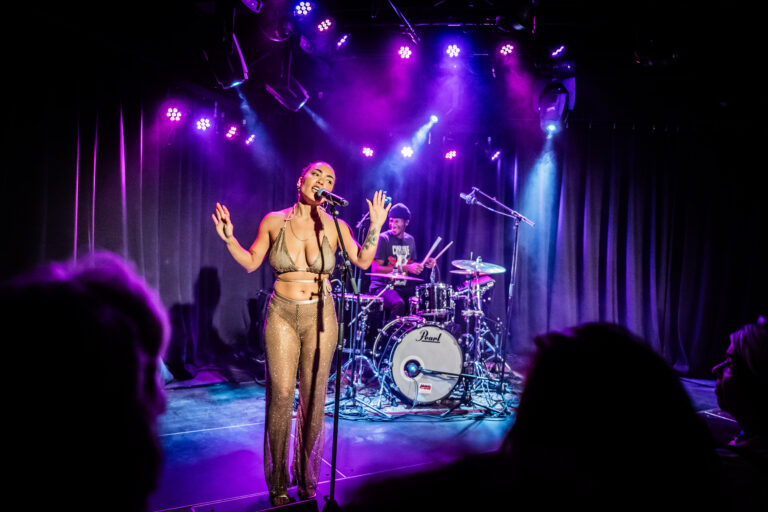 Een muzikante zingt op het podium terwijl een drummer achter haar speelt, omgeven door kleurrijke lichten.