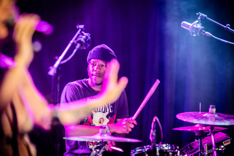 Een drummer in een donker speelstuk met paarse verlichting, terwijl hij geconcentreerd op zijn instrument speelt.