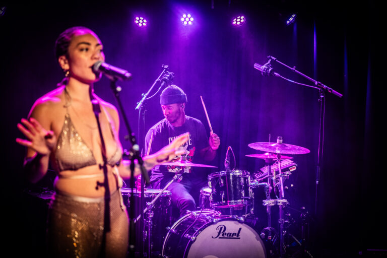 Een zangeres met een microfoon staat op het podium terwijl een drummer achter haar speelt, omgeven door paarse lichten.