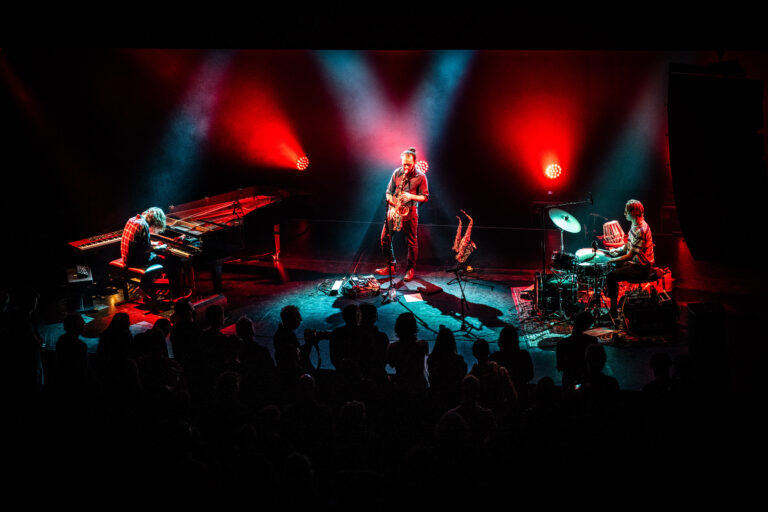 Een live band speelt op het podium met fel gekleurde lichten en een publiek dat toekijkt.