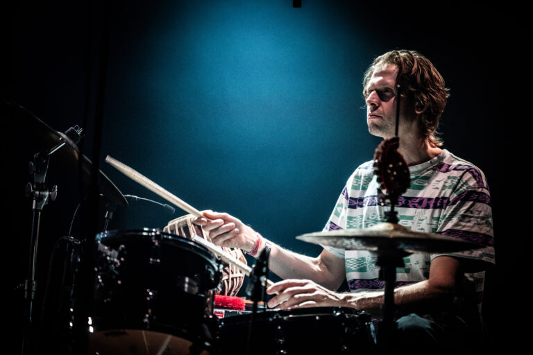 Een drummer speelt op een set tijdens een optreden, omringd door sfeerverlichting.