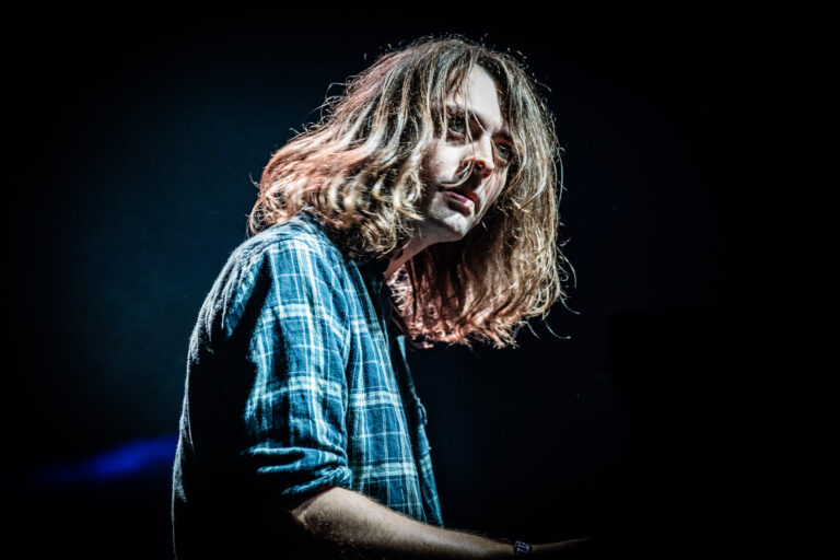 Jongen met lang, golvend haar, gekleed in een blauw-geruite blouse, kijkt geconcentreerd terwijl hij een instrument bespeelt op een podium.