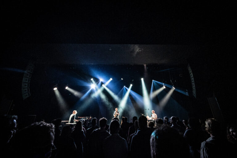 Een live muziekoptreden met verschillende muzikanten op het podium en een publiek dat naar het optreden kijkt.
