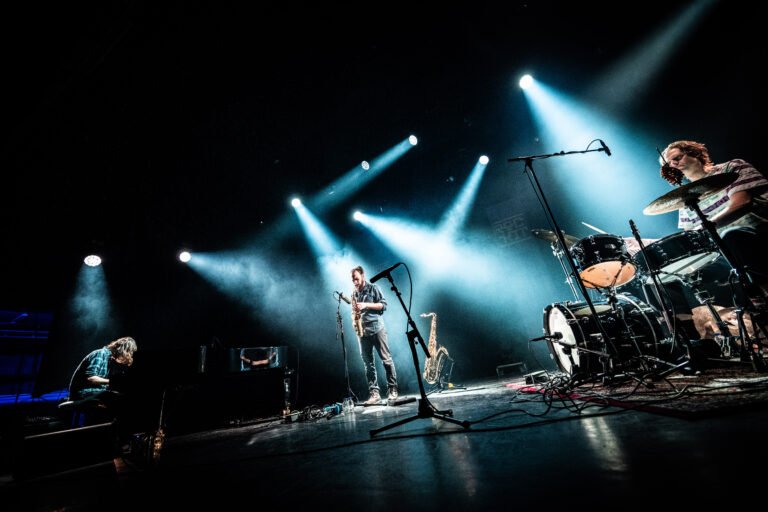 Een band staat op het podium met felblauwe lichten terwijl ze live muziek uitvoeren.