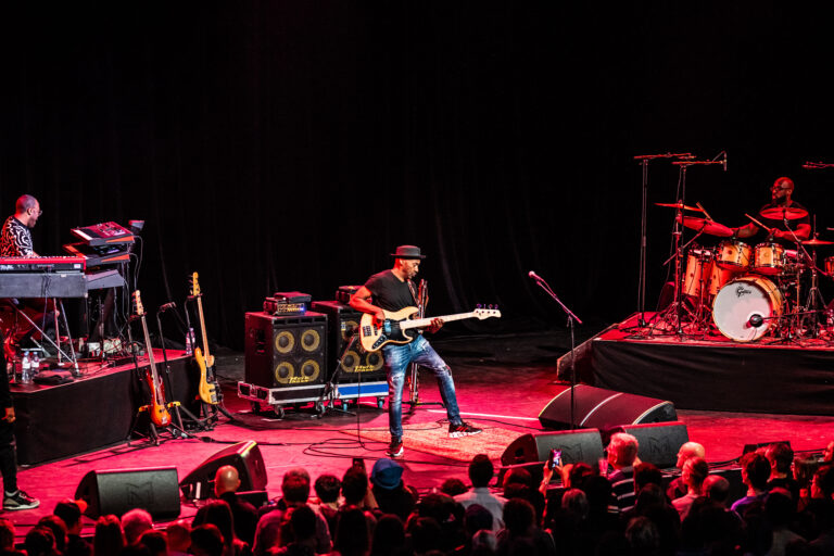 Een muzikant speelt gitaar op een live podium, omringd door apparatuur en een publiek.