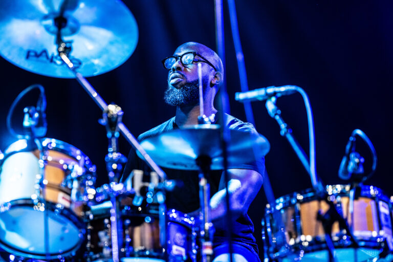 Een drummer speelt op een set met cymbals onder blauwe verlichting.
