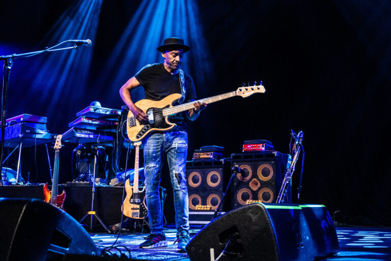 Een muzikant speelt gitaar op het podium met blauwe belichting en muziekapparatuur achter zich.