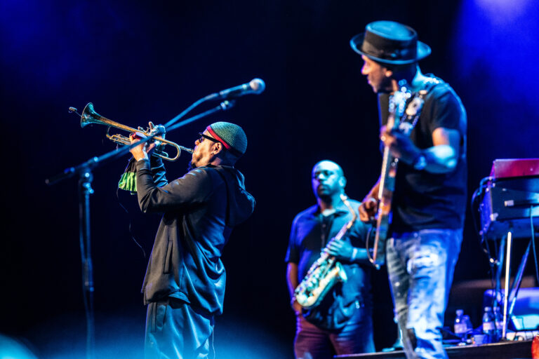 Een muzikant speelt trompet op een podium, terwijl twee andere bandleden luisteren en spelen.