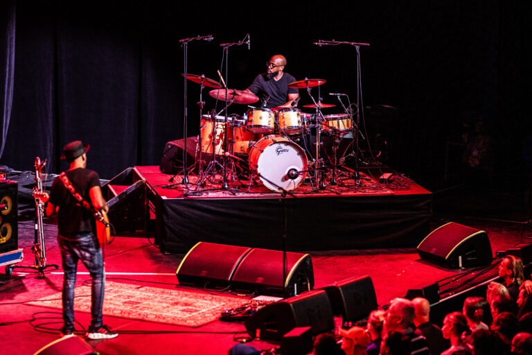 Een drummer speelt op een podium terwijl een gitarist in de spotlight staat, omringd door een publiek.