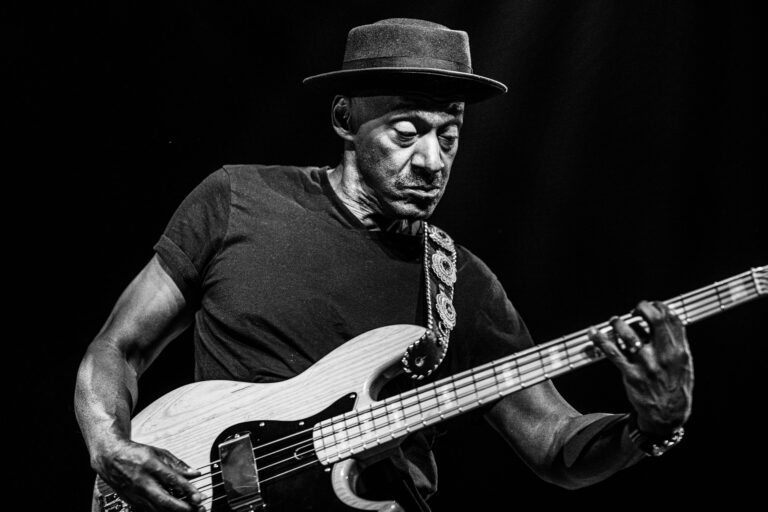 Een muzikant speelt gepassioneerd basgitaar op een podium, gekleed in een T-shirt en een hoed.