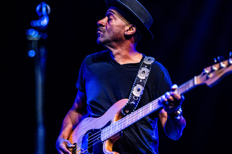 Een muzikant met een hoed speelt basgitaar op een podium in een sfeervolle setting.