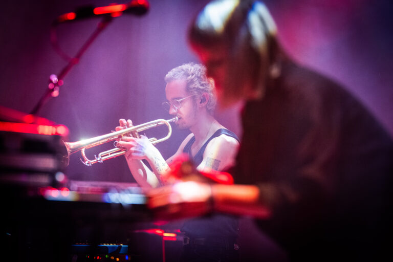 Een muzikant speelt trompet terwijl een andere muzikant aan een keyboard werkt, omringd door sfeerverlichting.
