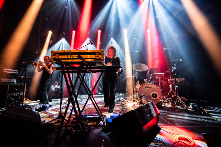 Een live optreden met een muzikant achter een keyboard, omringd door kleurrijke lichten en andere bandleden op het podium.