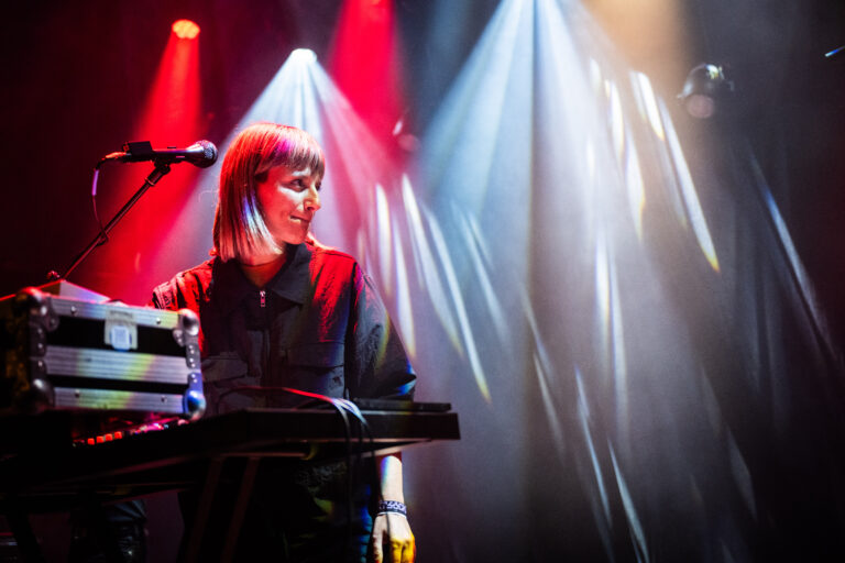 Een muzikante staat achter een synthesizer met kleurrijke verlichting op het podium.