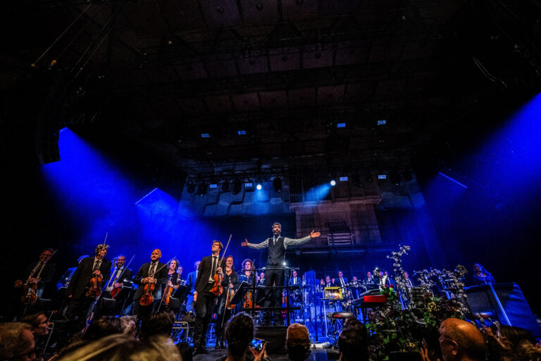 Een dirigent staat voor een orkest op een podium, omringd door instrumenten en kleurrijke lichten.