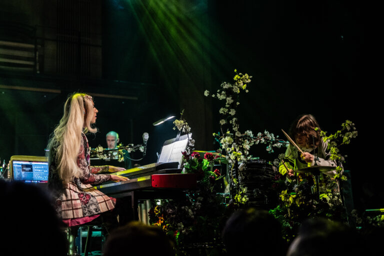 Een muzikante speelt piano op een podium dat is versierd met bloemen, terwijl een andere artiest op de achtergrond meedoet.