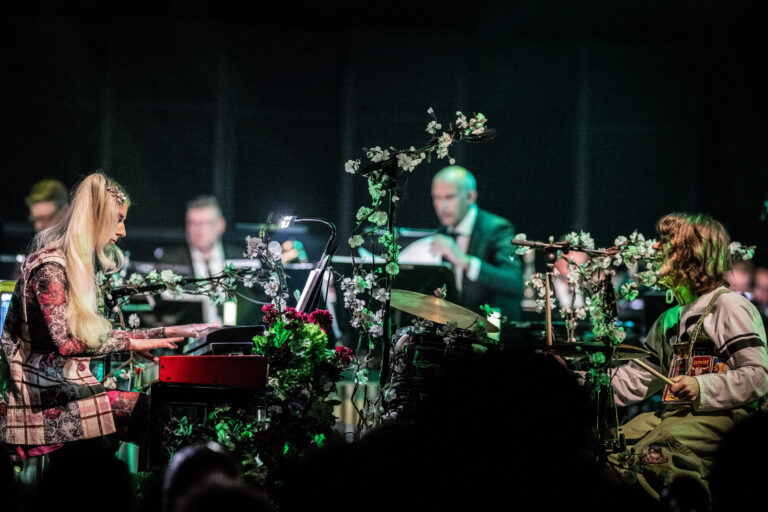Een groep muzikanten speelt op een podium, omringd door bloemen en groene decoraties.