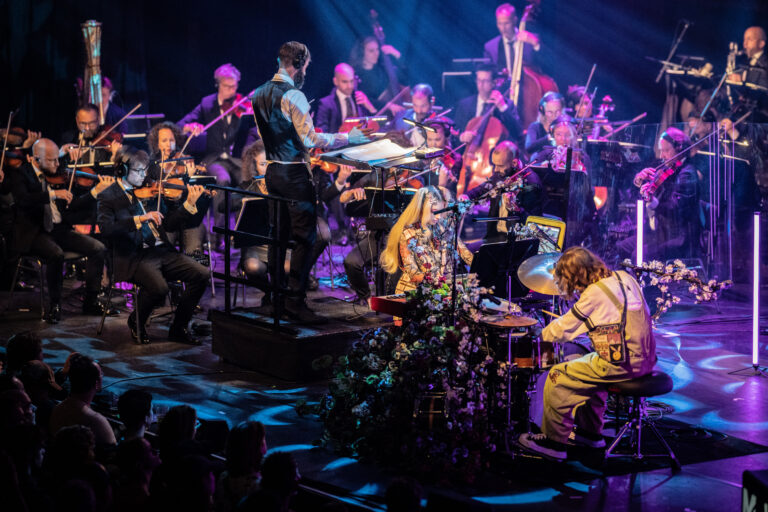 Een live orkest speelt op een podium, met een muzikant aan de drums en een dirigent die het optreden leidt.