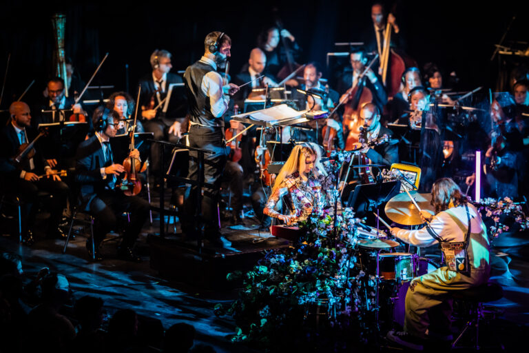 Een orkest met meerdere musici die optreden op een podium, omgeven door kleurrijke lichten en instrumenten.