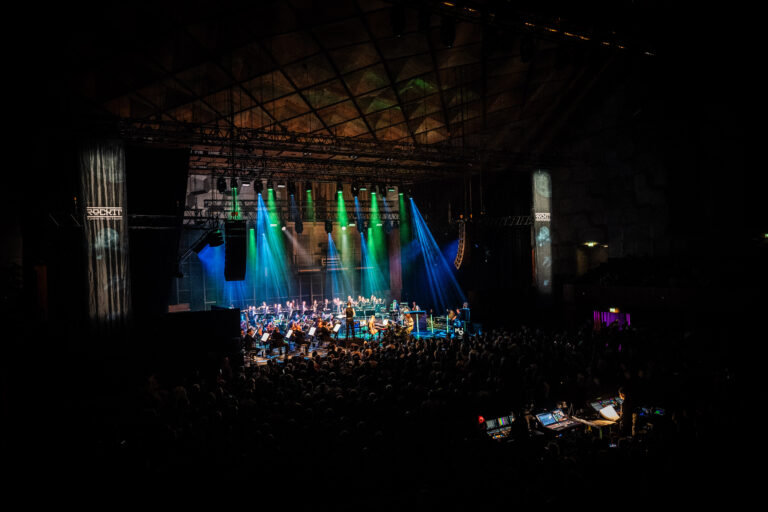 Een concert met een groot aantal muzikanten op het podium, verlicht door kleurrijke lichten.