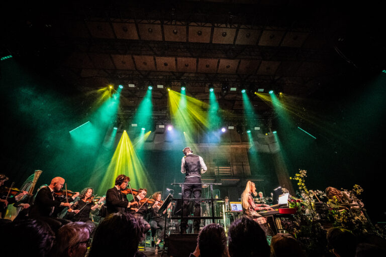 Een orkest speelt op een podium onder kleurrijke lichten, met een dirigent die leidt en muzikanten die spelen op verschillende instrumenten.