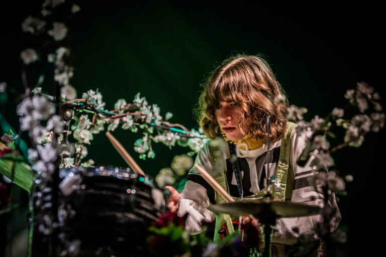 Een drummer speelt geconcentreerd op het podium, omringd door bloemdecoraties.