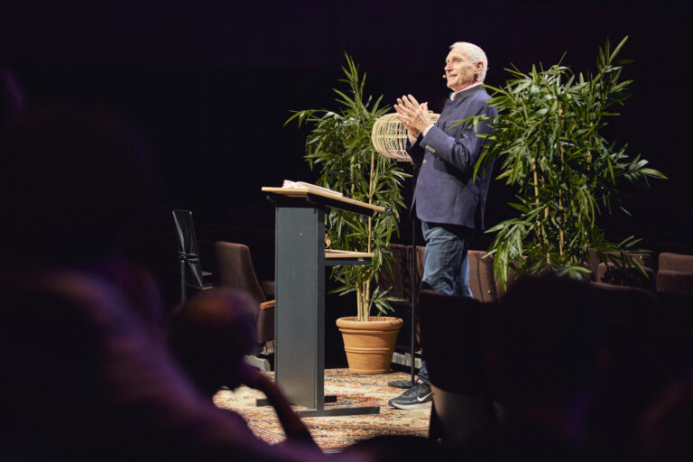Netjes geklede grijs harige man die spreekt voor een groep.