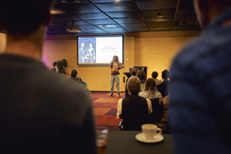 Vrouw die voor een groep mensen spreekt met een digibord op de achtergrond.