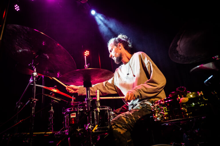 Drummer in actie op het podium, omringd door kleurrijke lichten.