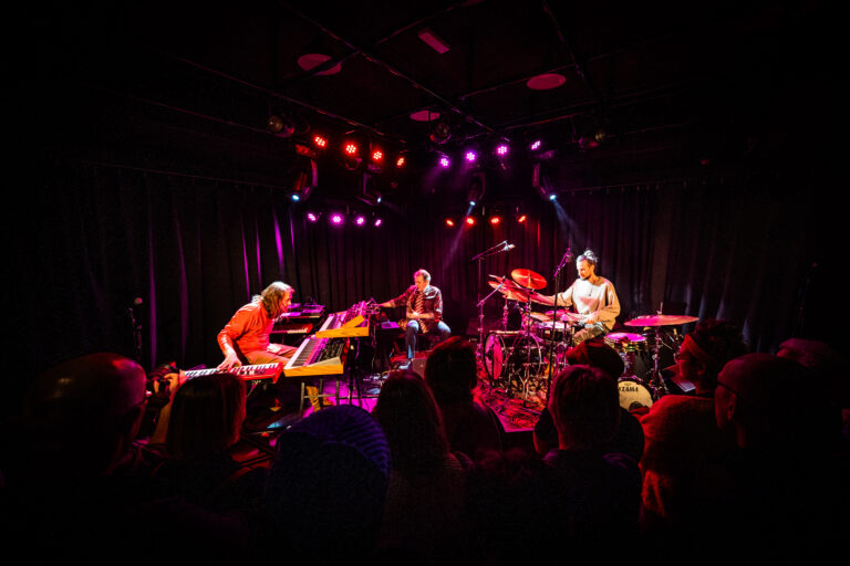 Drie muzikanten optreden op een podium met kleurrijke verlichting, terwijl het publiek kijkt.