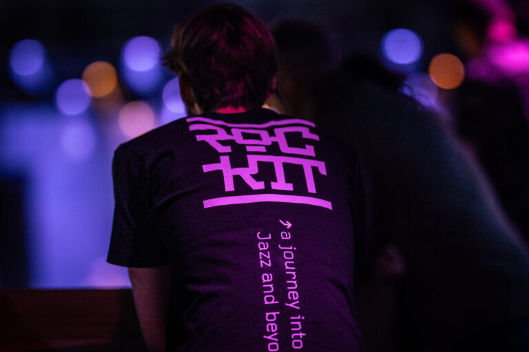 Een persoon met een zwart T-shirt met een wit en paarse opdruk, zittend met de rug naar de camera en een sfeervolle achtergrond.