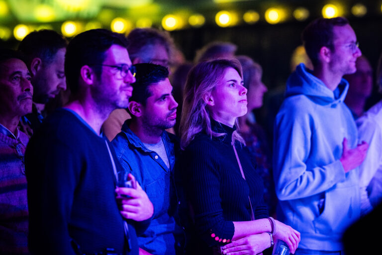 Een groep mensen kijkt aandachtig naar een podiumshow, omgeven door sfeervol verlichting.