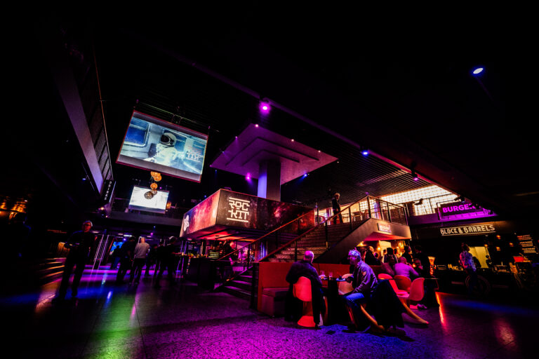 Een sfeervolle binnenruimte met kleurrijke verlichting en een moderne inrichting, waar mensen aan tafels zitten en een groot scherm is opgehangen.