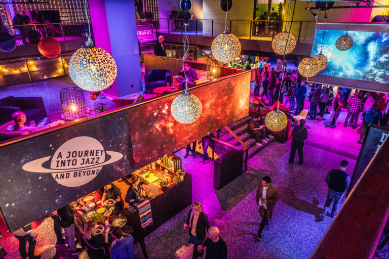 Een levendige evenementlocatie met kleurrijke verlichting en drukte van mensen, versierd met grote hangende lichten.