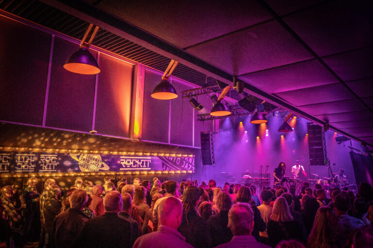 Een druk bezochte concertzaal met kleurrijke verlichting en een publiek dat naar het podium kijkt.