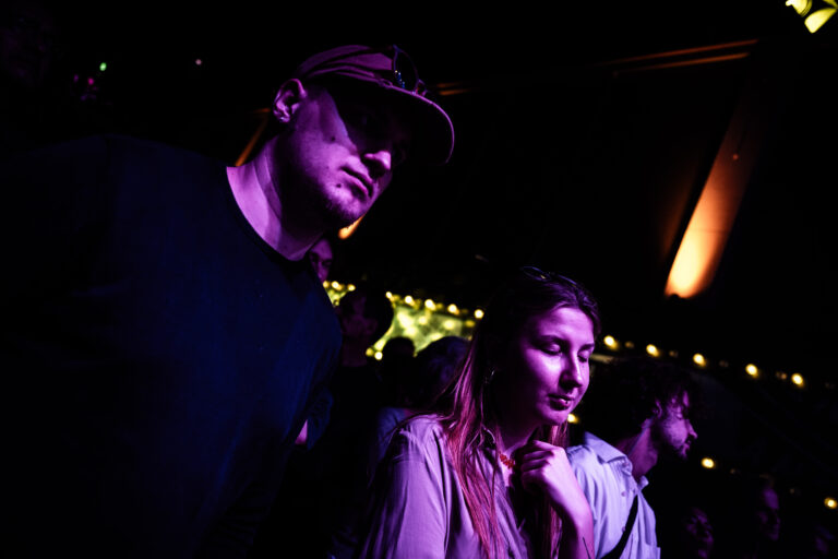 Een man en vrouw staan dicht bij elkaar in een donkere omgeving met kleurrijke verlichting, terwijl ze naar een optreden kijken.