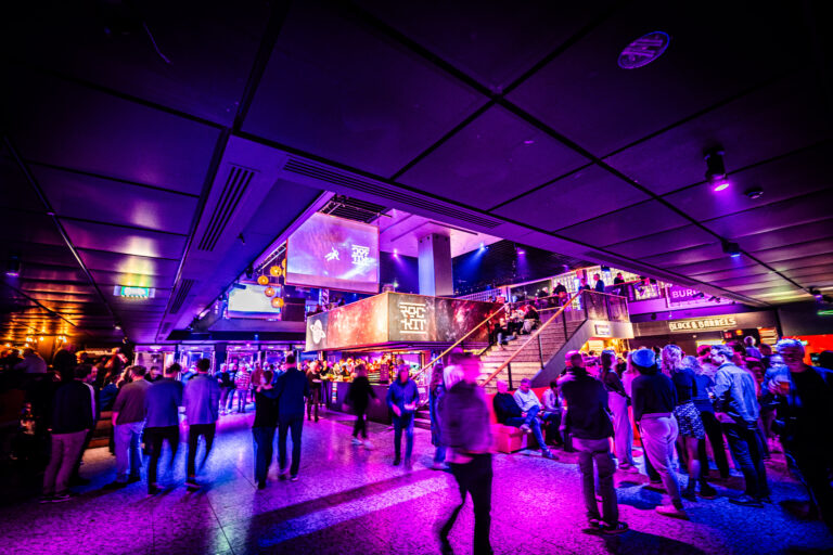 Een drukke, sfeervolle ruimte met kleurrijke verlichting en mensen die zich verplaatsen tussen verschillende etages.